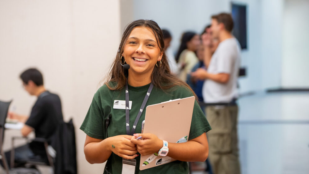 A young woman smiling in a green ASI Events polo with a clipboard and a communications headset