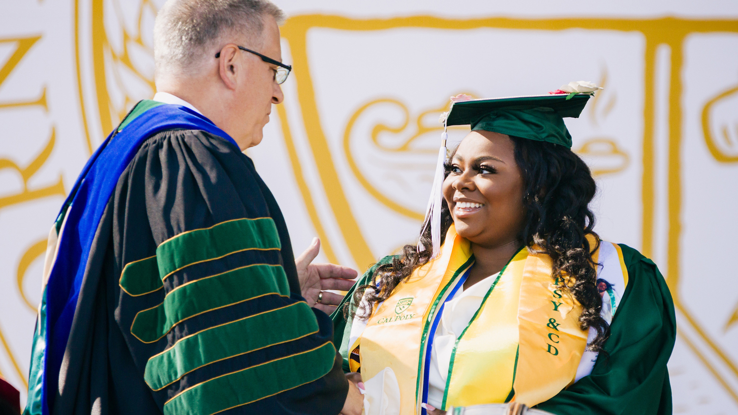 A college graduate in cap and gown shakes the hand of a university president in front of the Cal Poly seal