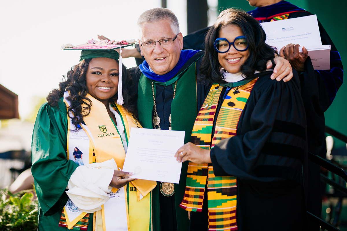 A student stands with a university president and a university supporter all wearing academic regalia at a Cal Poly commencement event