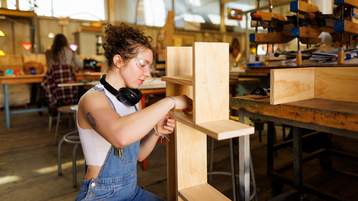 A student wearing overalls and safety glasses sands a wooden surface of a shelving unit in a fabrication shop