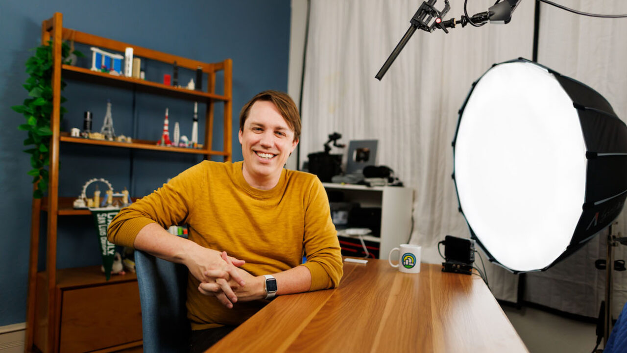 A man sits at a desk in a recording studio decorated with city-themed Legos, with a microphone and light box pointed at him