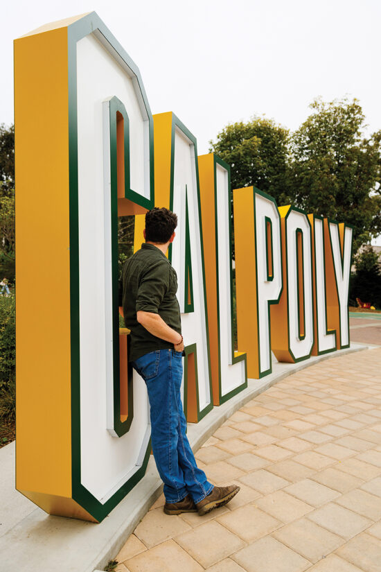 A man in a green shirt and jeans leans against a large 3-D Cal Poly sign.