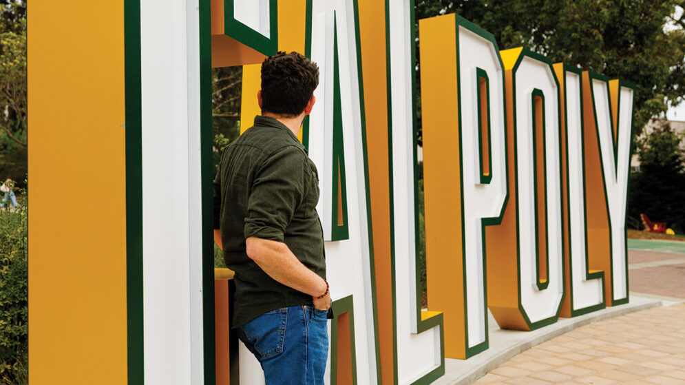 A man in a green shirt and jeans leans against a large 3-D Cal Poly sign.