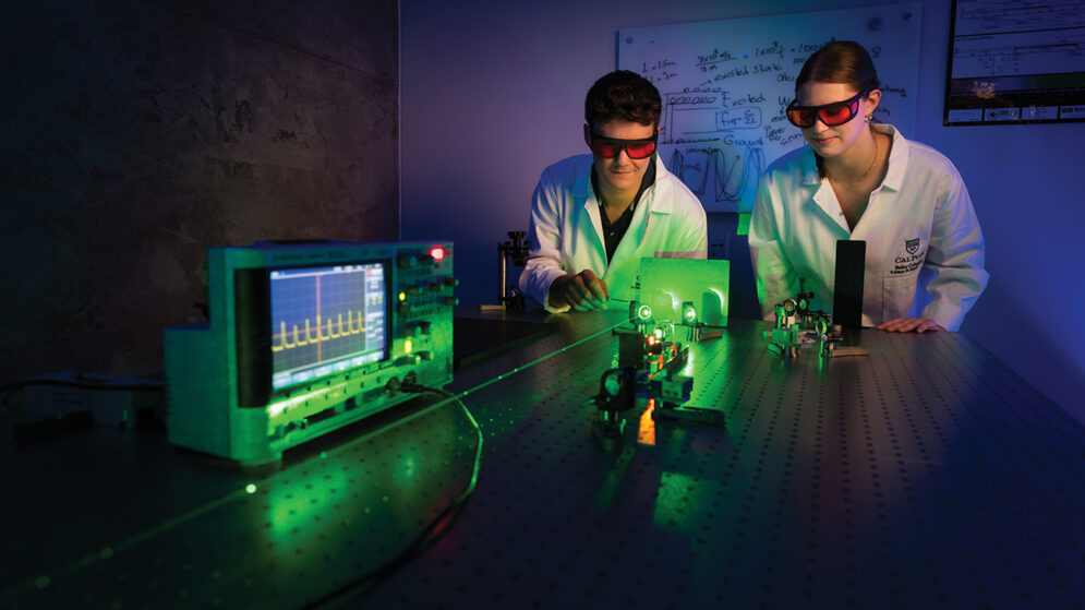 In a dark lab, two students adjust a device projecting a green laser beam across a table mounted with scientific instruments.
