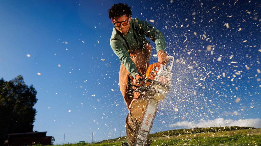 A man in sunglasses stands on a log as he cuts the end off with a chainsaw