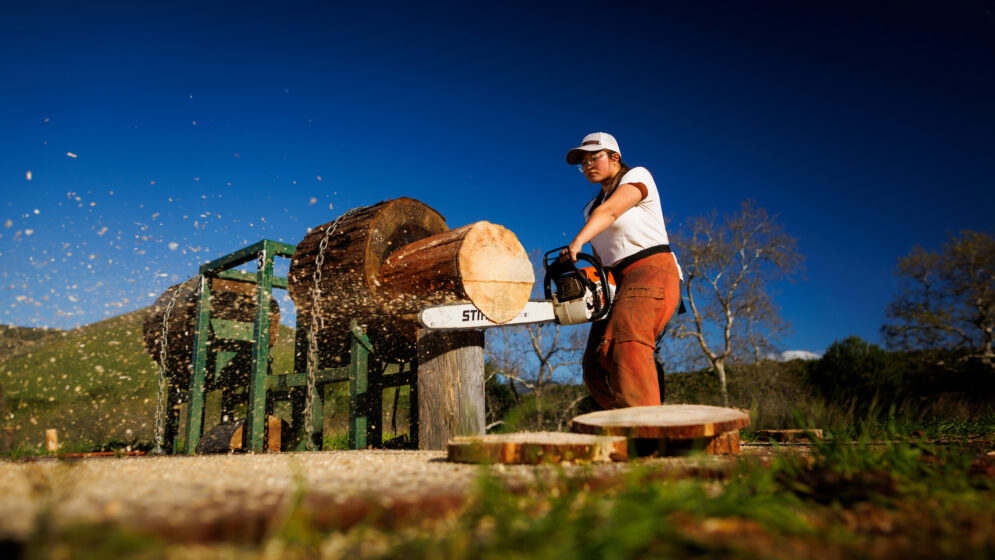A young woman in a baseball cap and safety glasses slices the end off a large log with a chainsaw