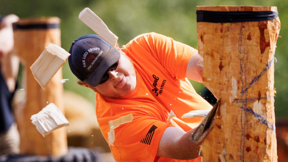 A man in an orange shirt and baseball cap sends chunks of wood flying as he chops a vertical log with an axe