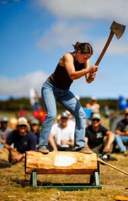 A young woman swings an axe down to chop a log she is standing on