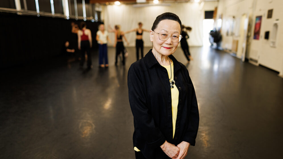 A woman in a black jacket smiles in front of a dance studio with dancers rehearsing in the background.