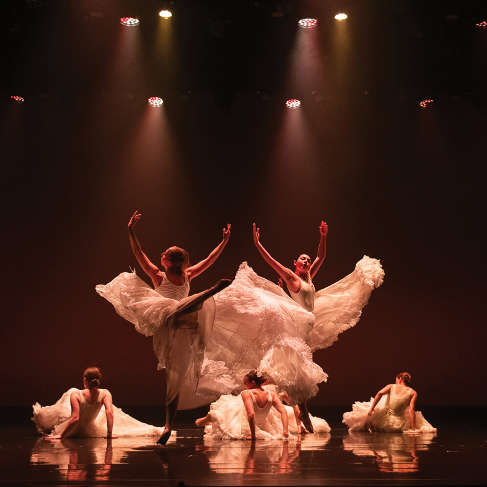 Five dancers in white dresses dance under lights on stage