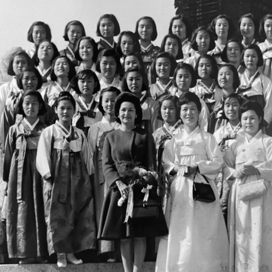 In a black and white vintage photo, a group of girls in traditional Korean dresses pose behind three older women.