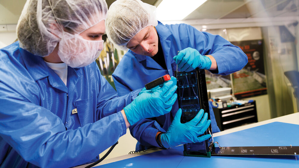 Two students in lab gear work on a rectangular black device in a lab