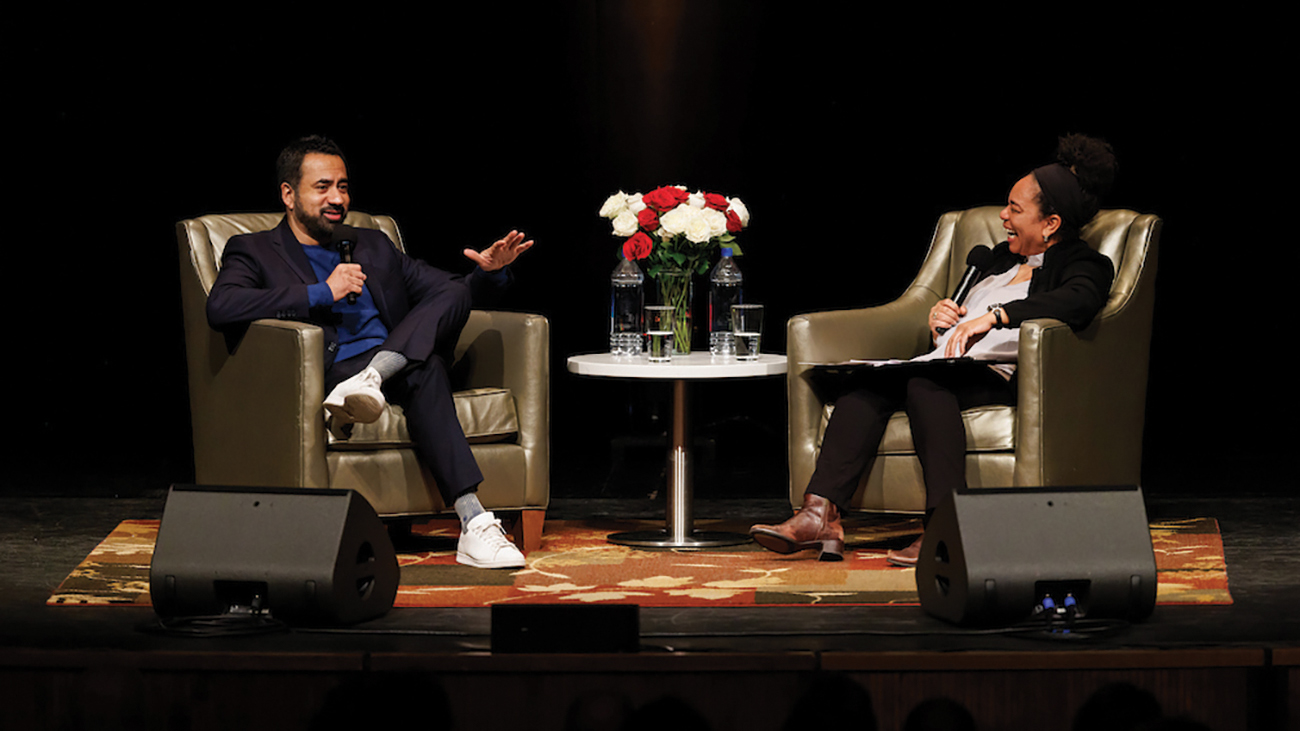 A man and woman seated in chairs facing each other on a stage speak into microphones