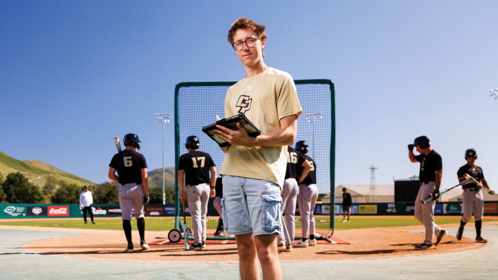 A student assistant stands with a tablet near the Cal Poly Baseball team practice