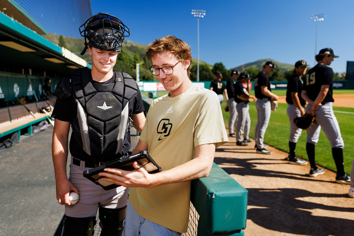 A baseball player wearing catchers gear stands next to a student assistant with a tablet in the dugout while other players line up on the foul line