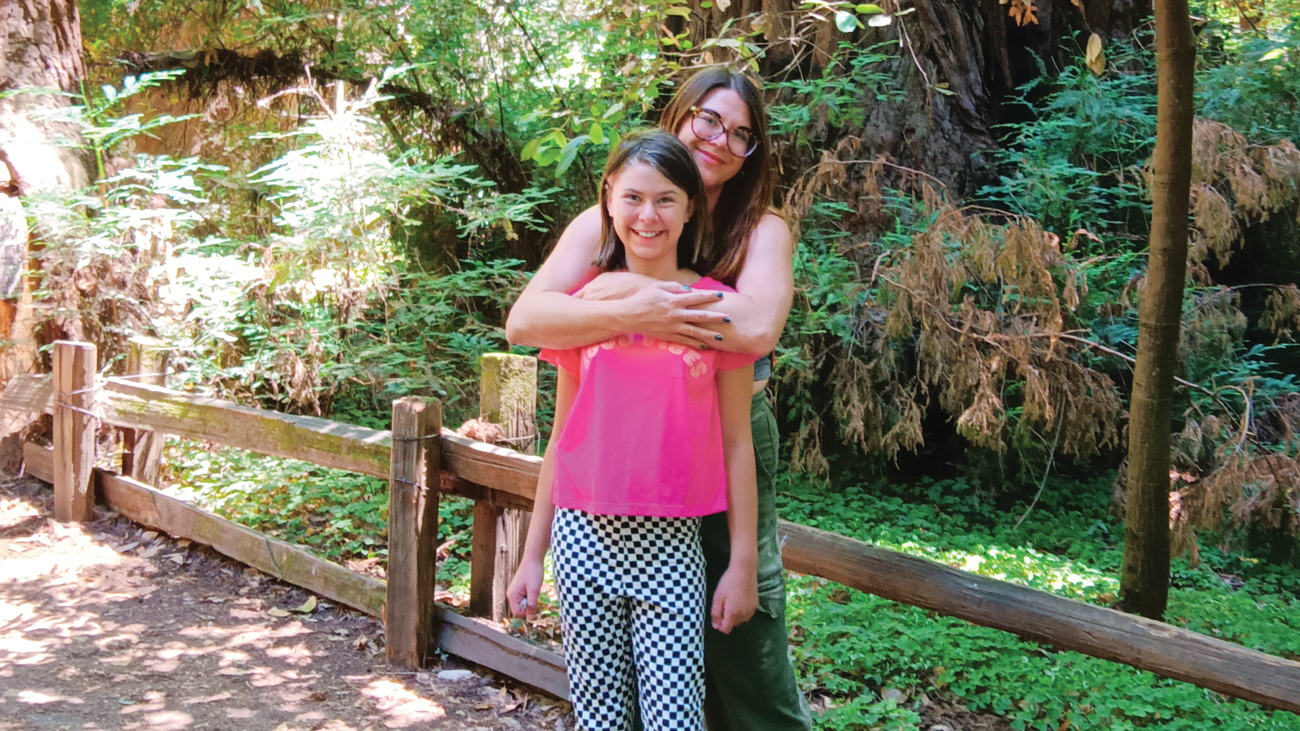 A mother and daughter embrace near a grove of large trees