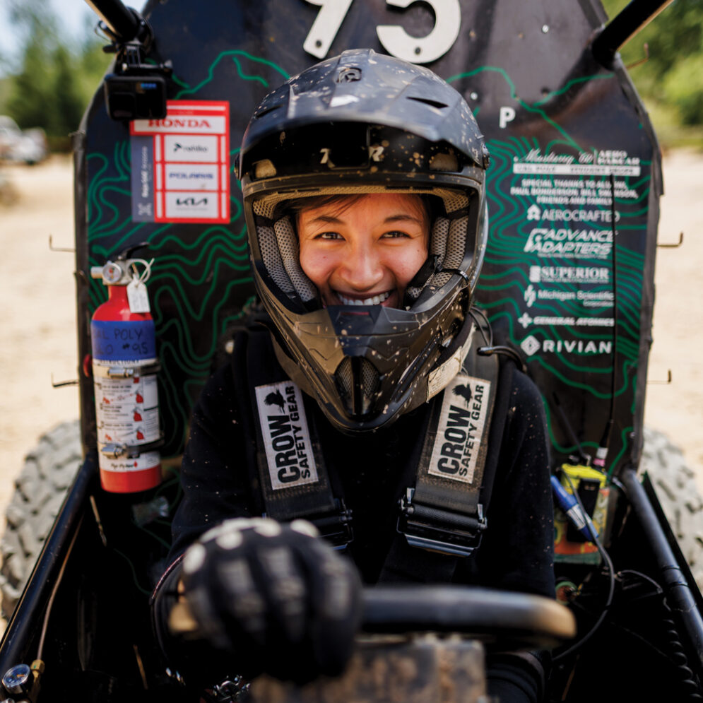 A Cal Poly Racing driver smiles while wearing a helmet and strapped into the driver's seat of an off-road vehicle