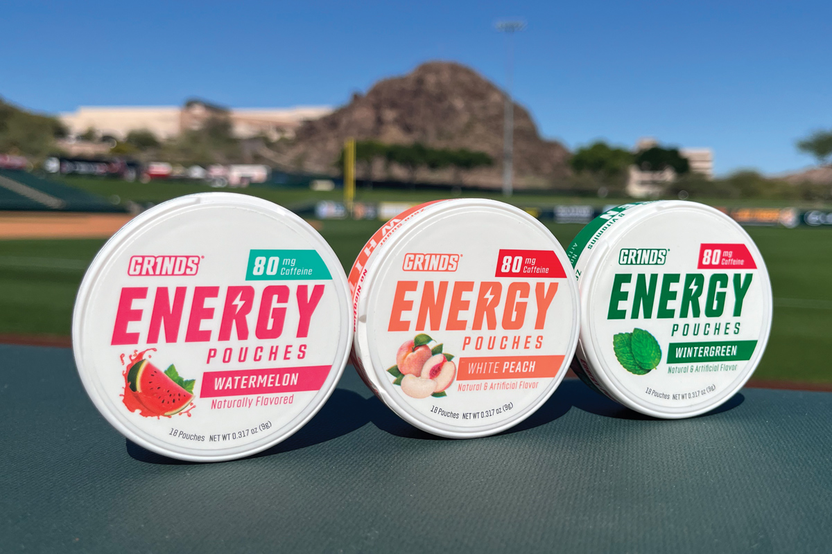 Three packages of Grinds Pouches displayed outdoors with an athletic field and a mountain behind them