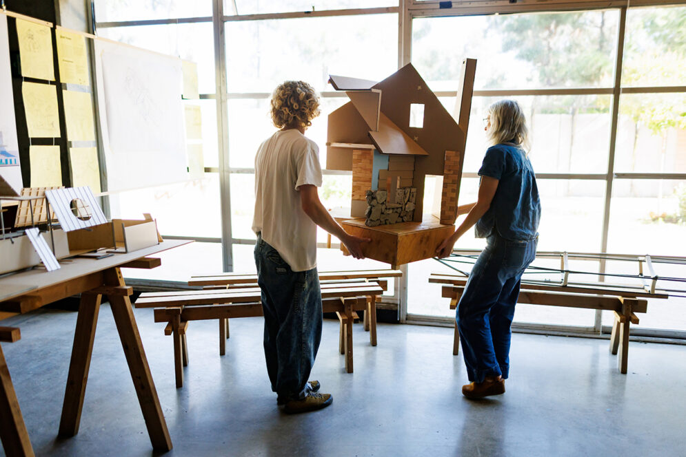 A young man and a woman carry an architectural model of a house toward a display table.