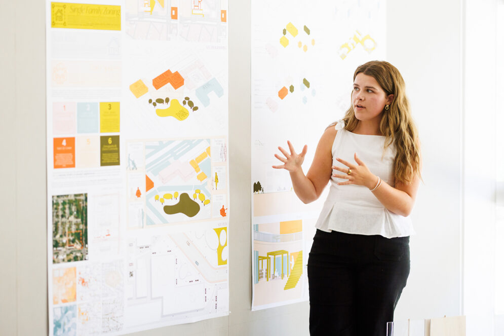 A young woman gestures toward a large poster display of colorful architectural renderings