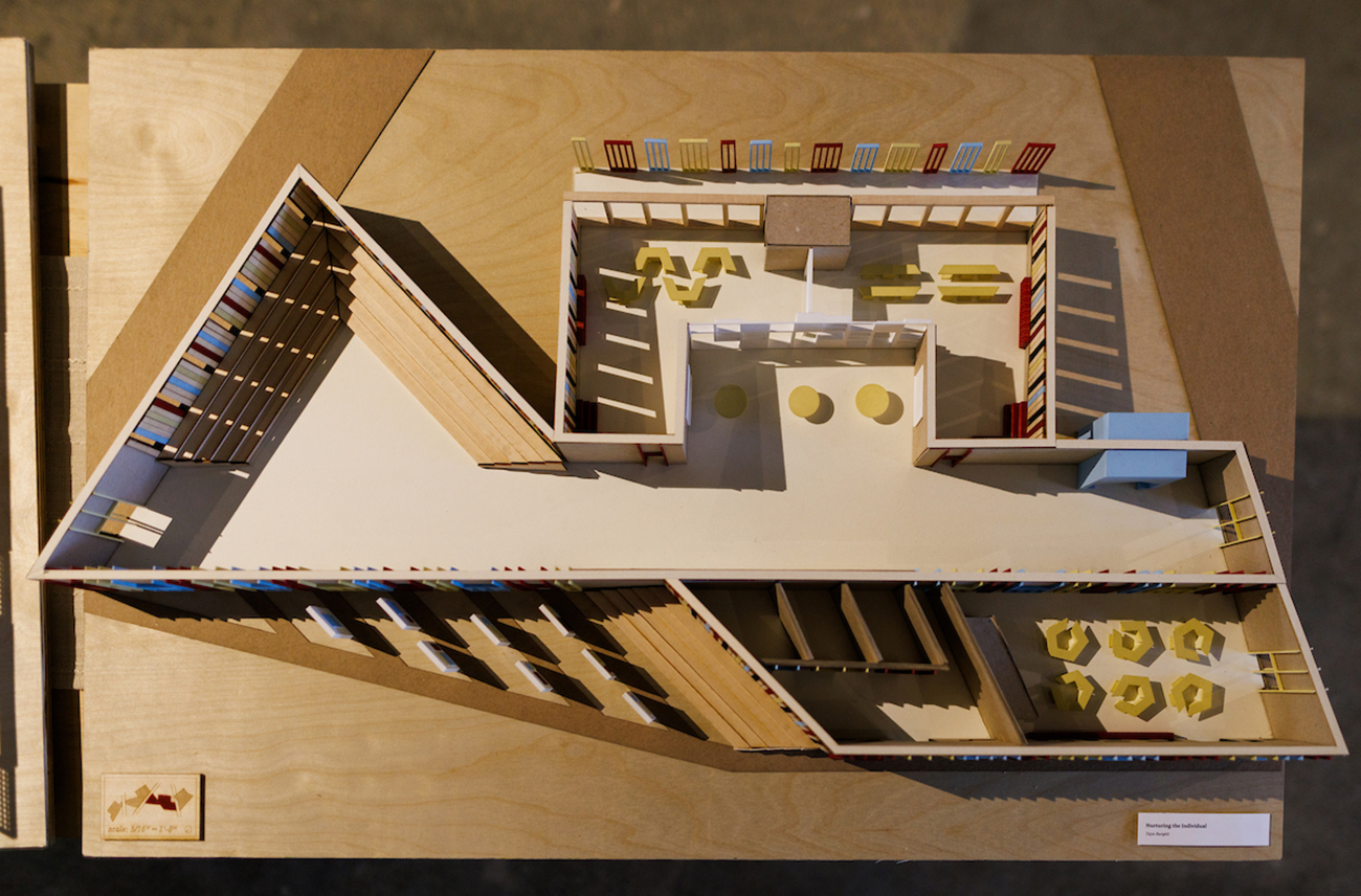 From above, an angular architectural model of a building casts a shadow across a wooden display table.