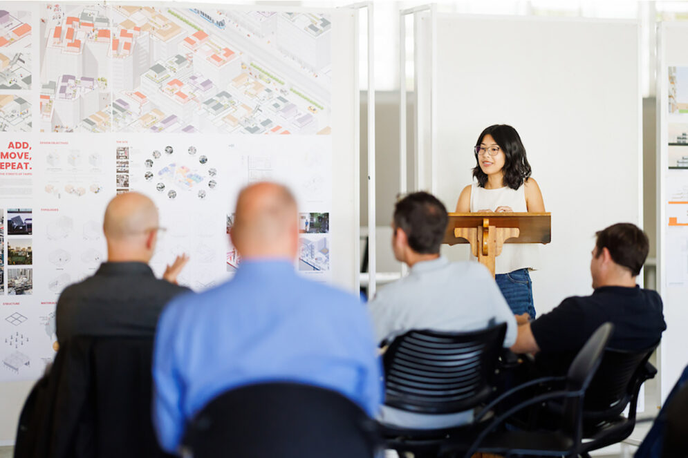 A young woman stands at a podium giving a presentation to a group of older professionals next to a colorful poster display