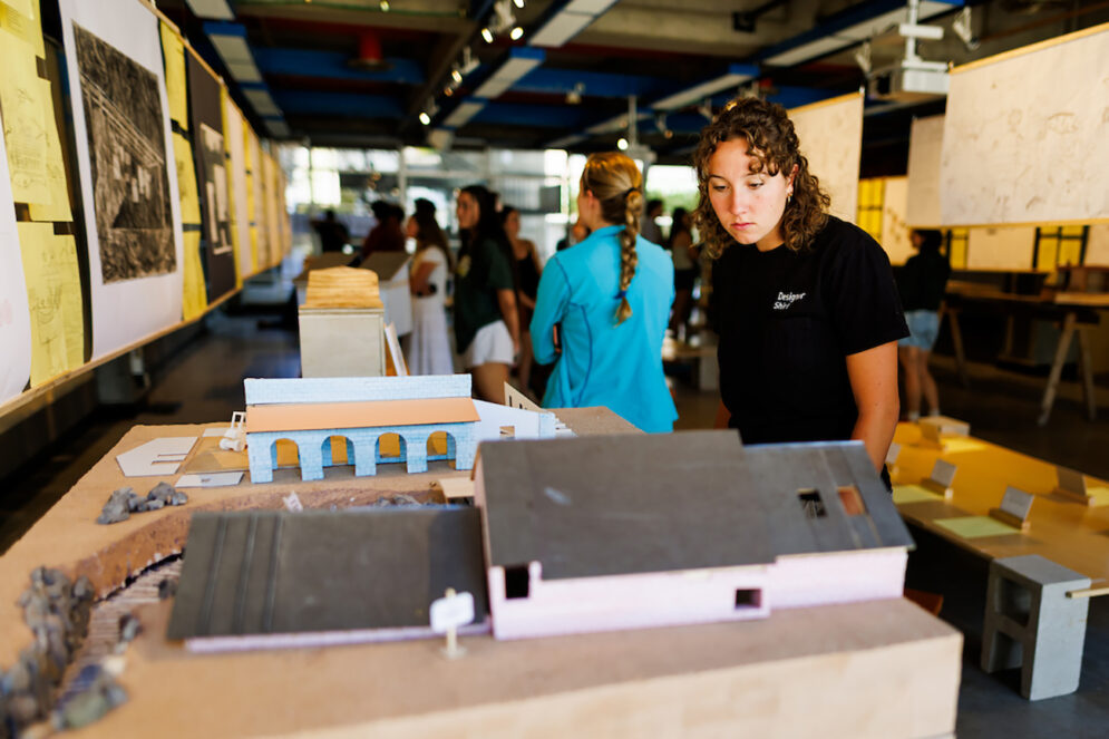 A young woman looks at a model of a building in a crowded room full of architectural displays.