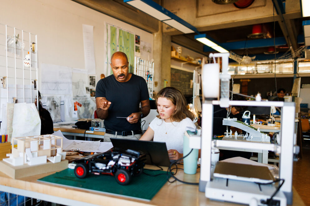 In a lab filled with architectural models, a professor guides a young woman working on a computer.