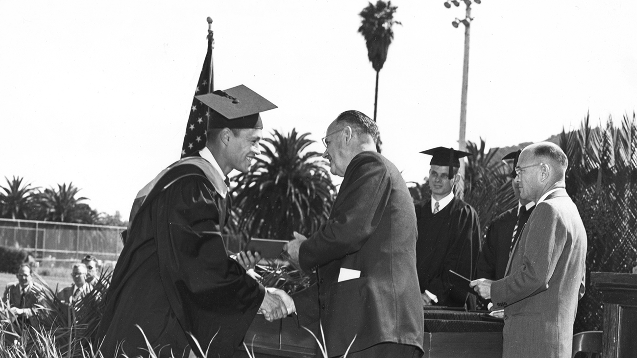 A man in a suit shakes hands with a young man in a graduation cap and gown in a vintage black and white photo