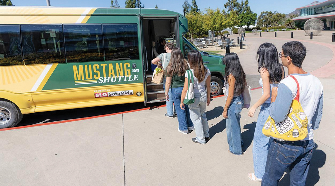 A group of students line up to enter a green and gold bus at a campus bus stop.