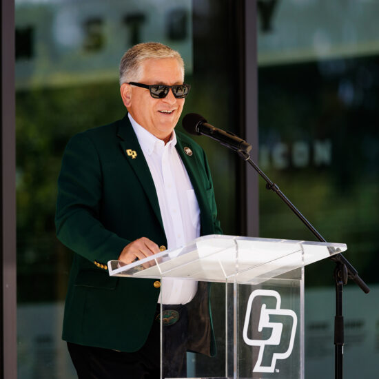 A man in a green blazer and sunglasses speaks in front of a podium marked with CP