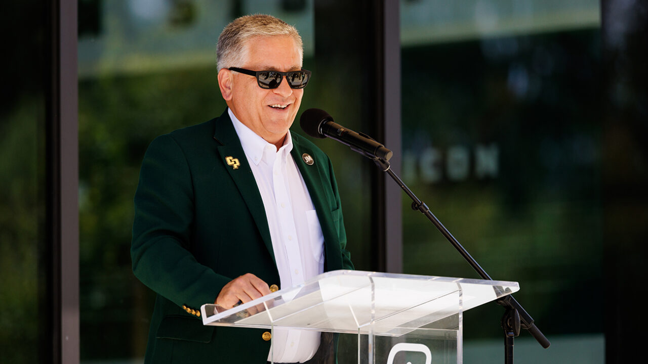 A man in a green blazer and sunglasses speaks at a podium marked with CP