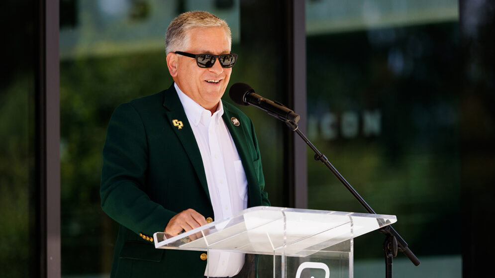 A man in a green blazer and sunglasses speaks at a podium marked with CP