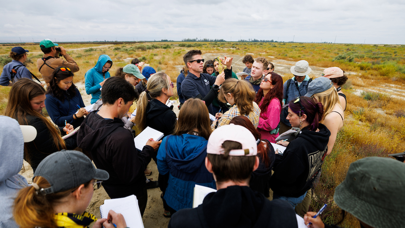 Professor Matt Ritter holds a plant up amid a group of students with notebooks during a field biology trip.