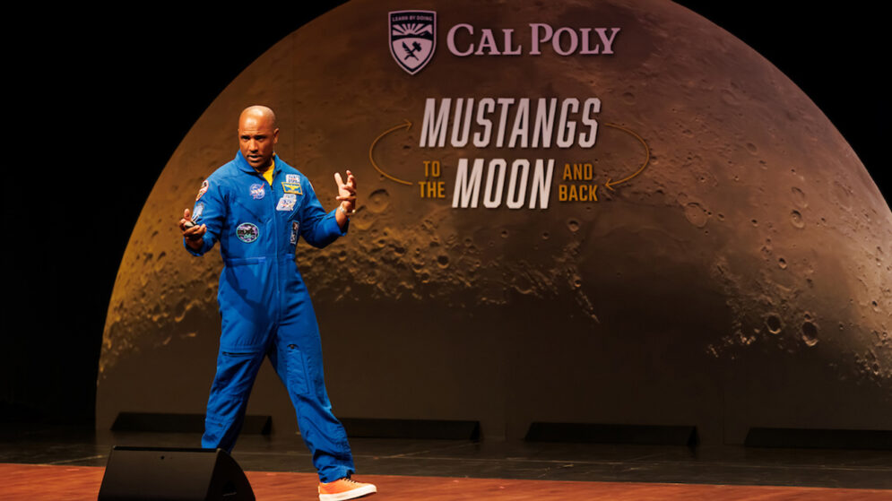 A man in a blue NASA flight suit speaks on stage in front of an image of the moon and the words "Mustangs to the Moon and Back"