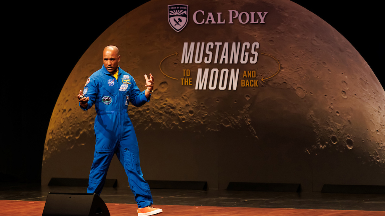 A man in a blue NASA flight suit speaks on stage in front of an image of the moon and the words "Mustangs to the Moon and Back"