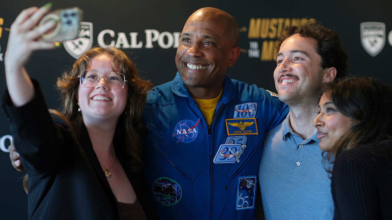 A man in a blue NASA flight suit poses for a photo with a group of Cal Poly students.