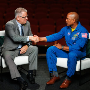 A man in a grey suit and a man in a blue NASA flight suit shake hands