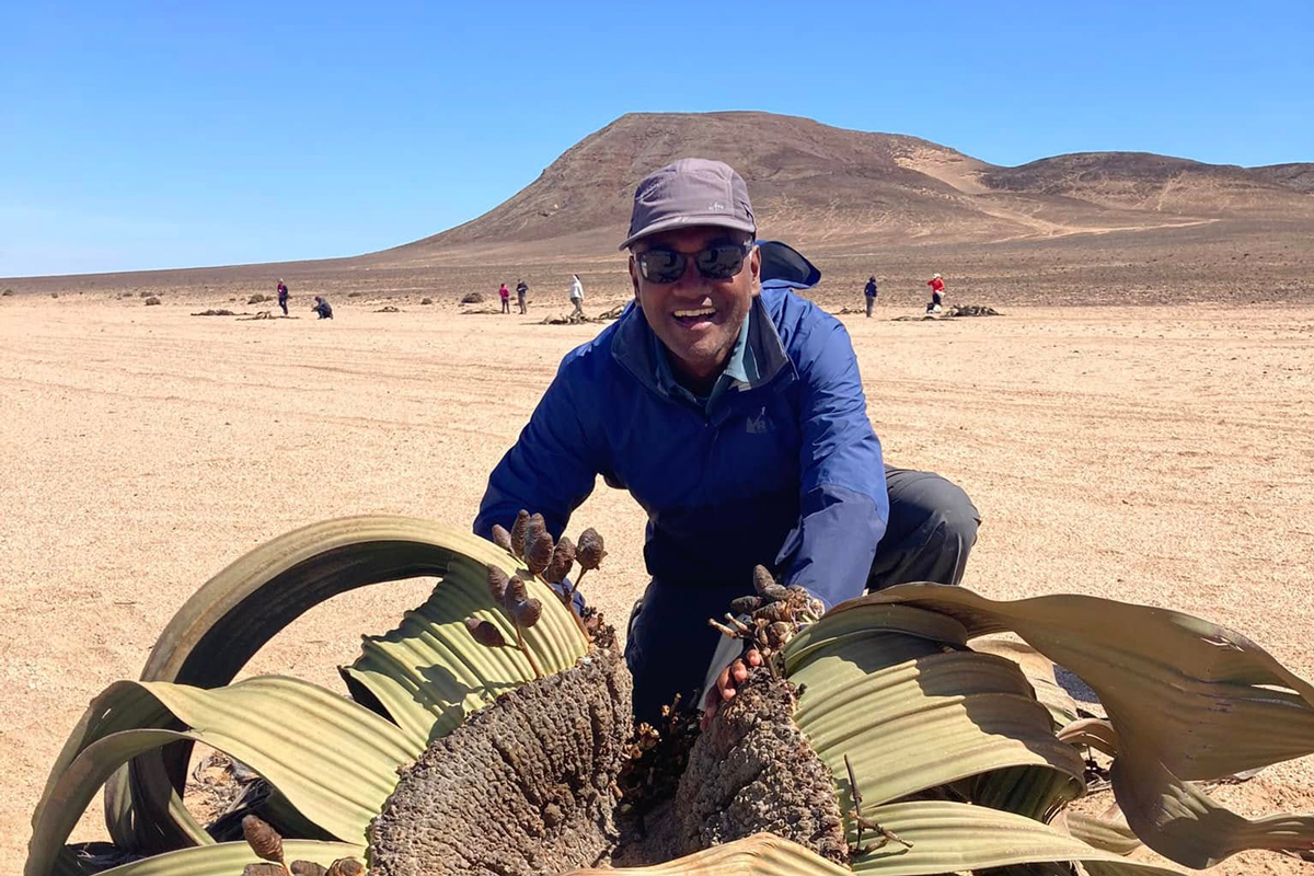 A biologist smiles while kneeling next to a rare plant in the Namib Desert.