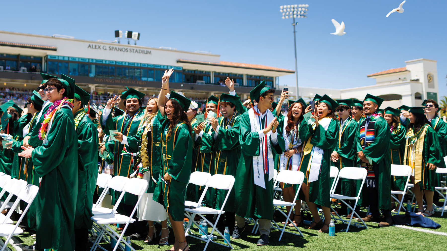 Cal Poly graduates in green cap and gown stand in Spanos Stadium with doves flying overhead