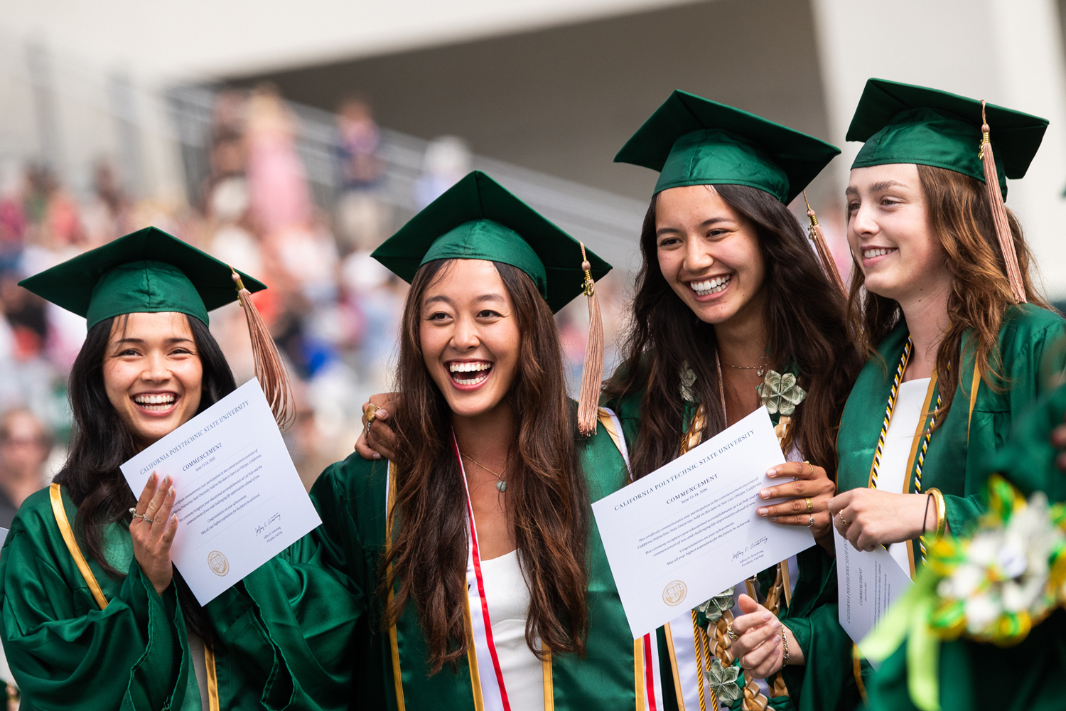 Four graduates in green caps and gowns smile during Cal Poly commencement