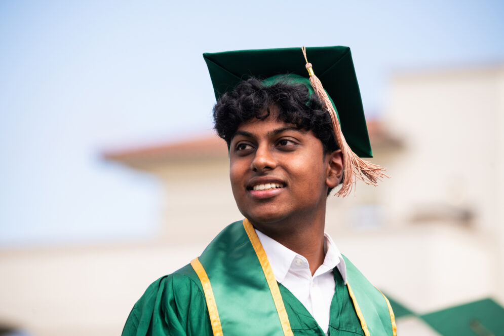 A Cal Poly graduate in green cap and gown looks toward the stands during commencement ceremonies