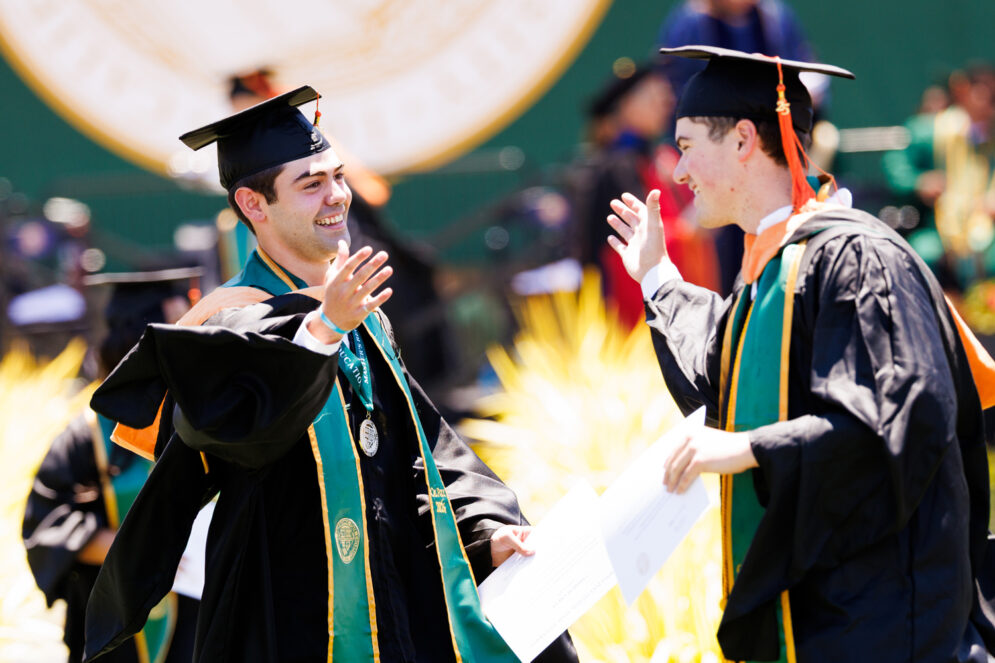 Two master's program graduates in black robes high five after crossing the commencement stage.