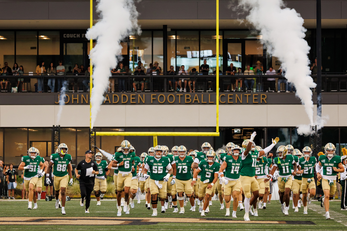 The Cal Poly football team wears green and gold uniforms while taking the field in front of the John Madden Football Center