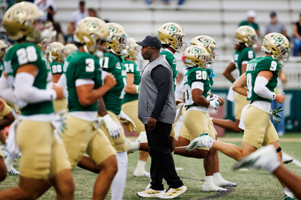 Cal Poly Football Head Coach Tim Skipper stands amidst uniformed team members as they warm up.