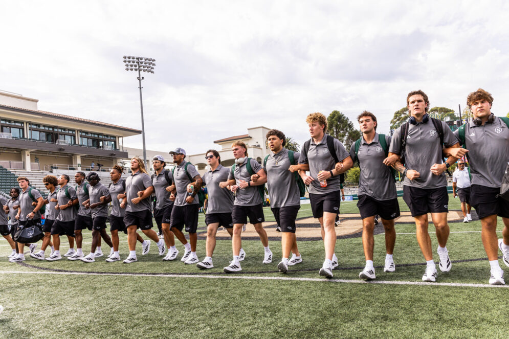 The Cal Poly football team members link arms while walking across Mustang Memorial Field in Spanos Stadium.