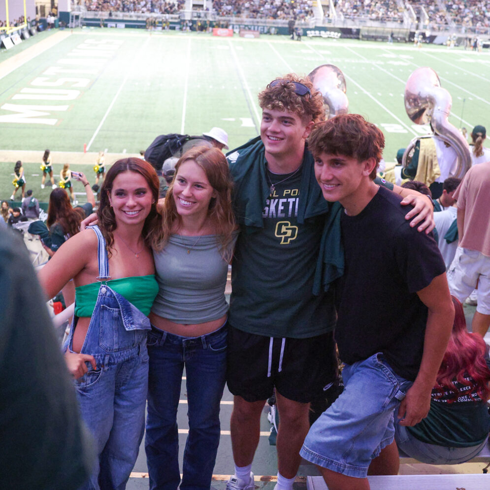 Four students pose for a photo in the student section of the Spanos Stadium stands during a Cal Poly football game.