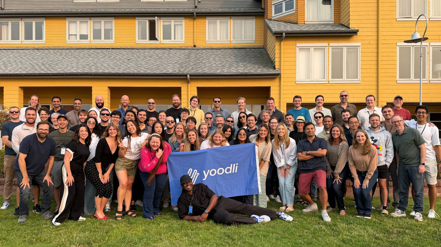 Dozens of people smile standing on grass with a blue Yoodli banner