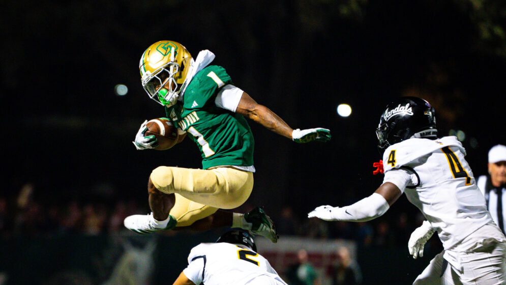 A Cal Poly football player holds a football and jumps over defenders during a football game in Spanos Stadium.
