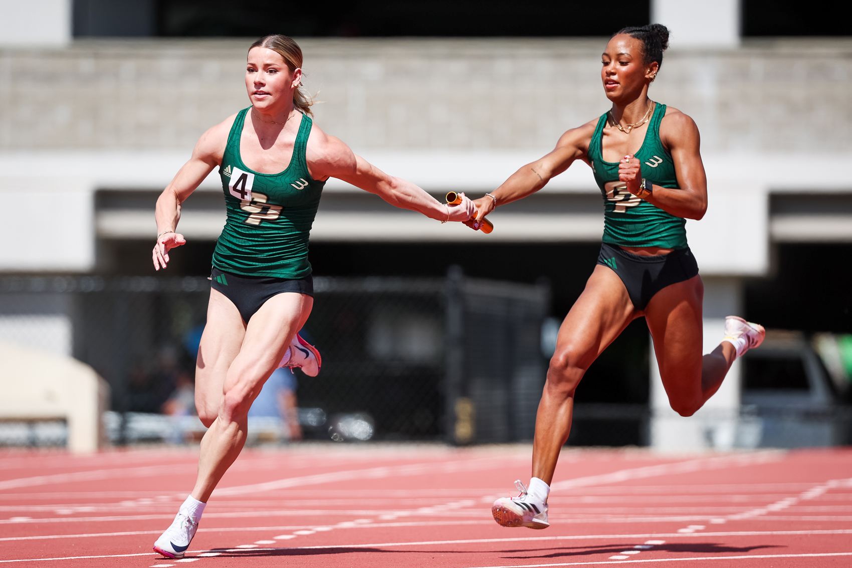 A track athlete passes a baton to another while wearing Cal Poly uniforms